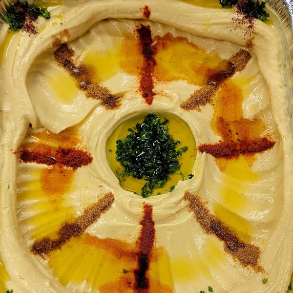 Creamy hummus with a swirl of olive oil, parsley, and spices in a foil container.