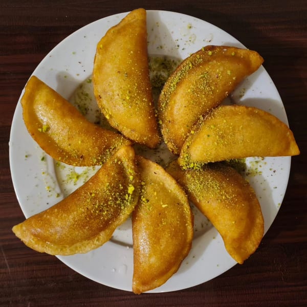 Seven golden-brown, crescent-shaped Mediterranean pastries, sprinkled with green pistachios, arranged on a white plate.