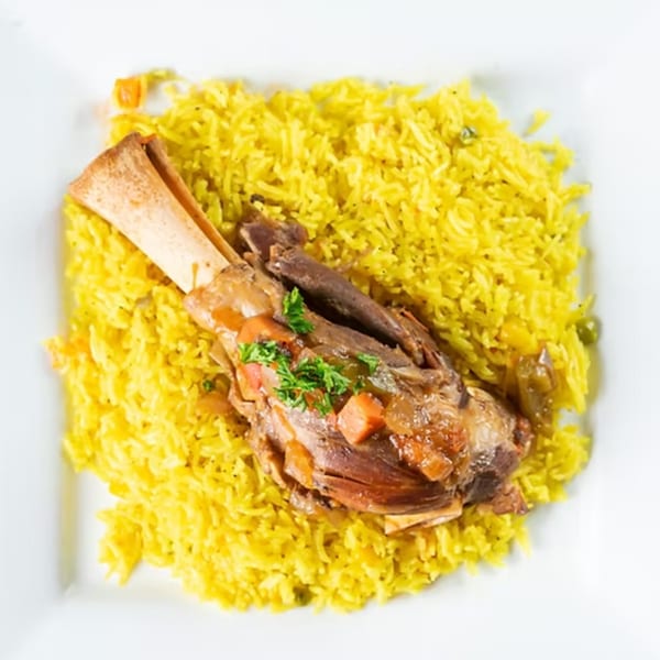 Braised lamb shank with bone, garnished with parsley, served over yellow rice on a white plate.